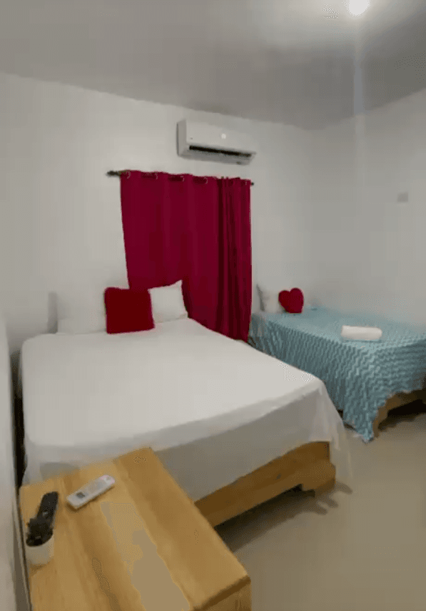 Dormitorio con cama blanca, cortinas rojas, aire acondicionado y cama individual con colcha azul.