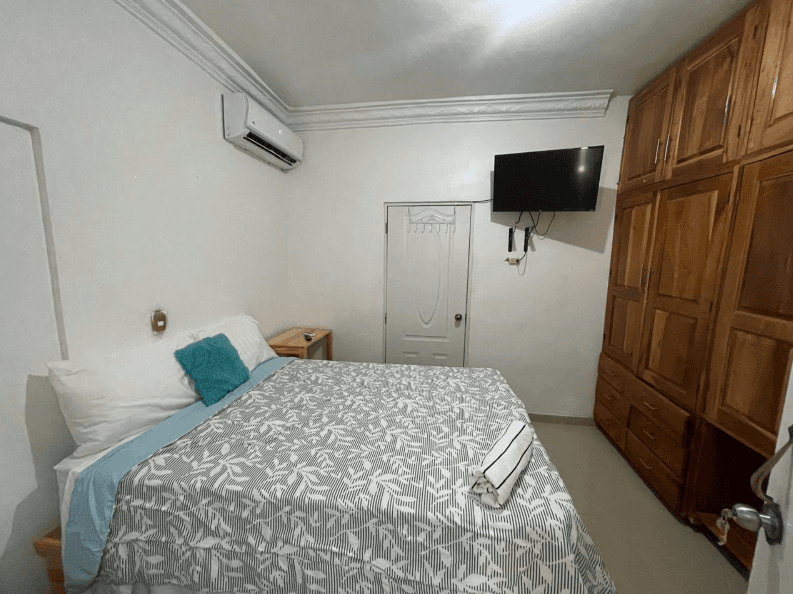 Dormitorio con cama de edredón estampado, televisor en pared, aire acondicionado y armario de madera.