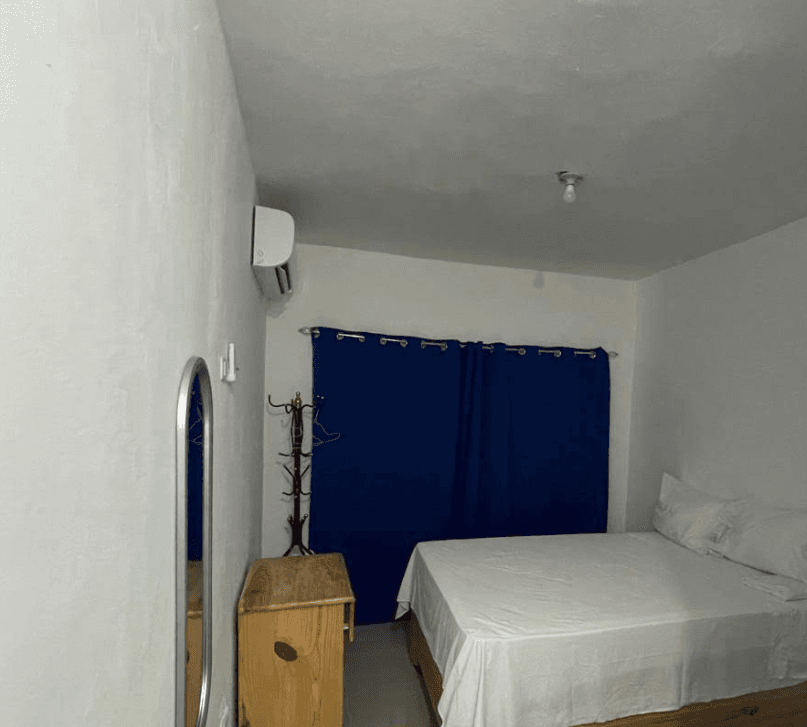 Habitación blanca con cama, cortinas azules, perchero de madera y aire acondicionado en la pared.