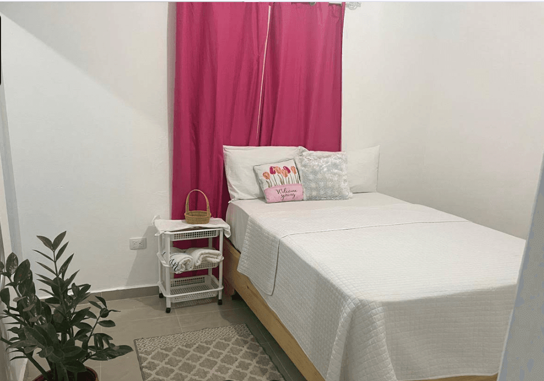 Cama blanca con cojín de primavera, cortinas fucsia, planta verde y carrito con toallas.