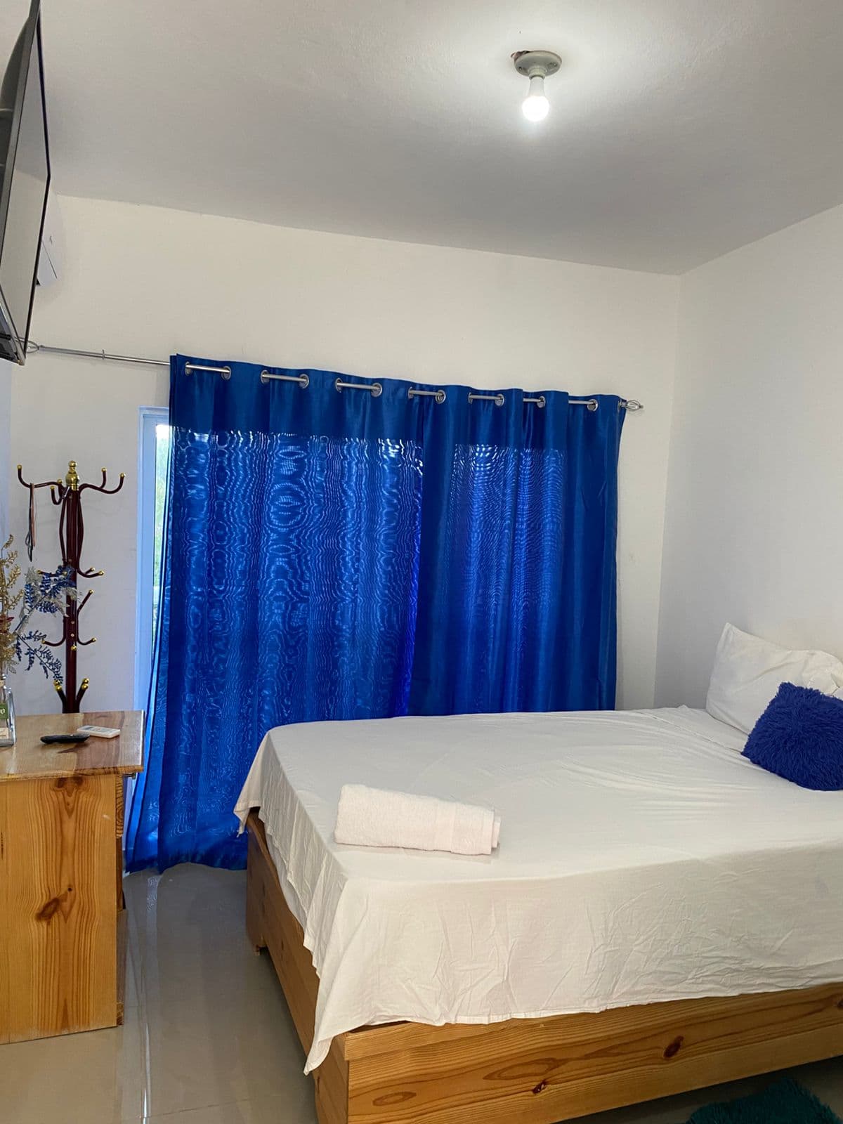 Dormitorio con cama blanca, cortinas azules brillantes, mesita de madera y paredes blancas.