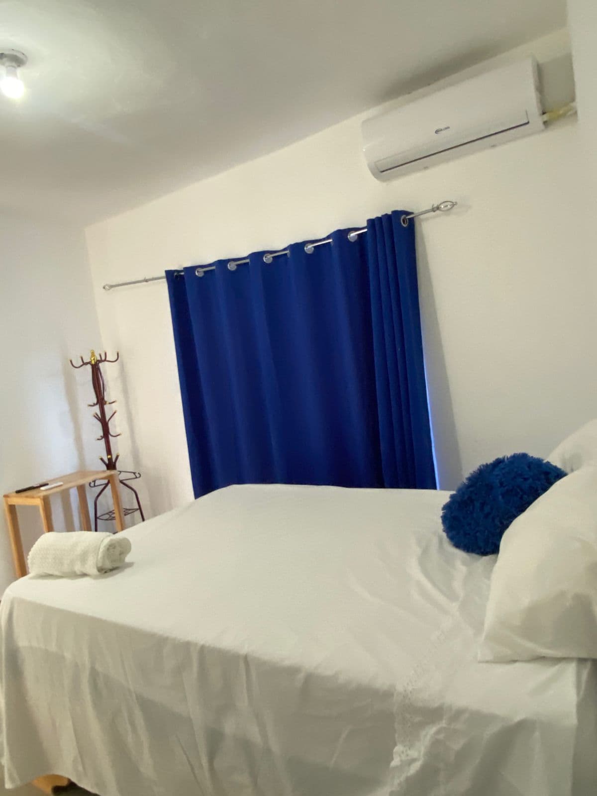Cama blanca con almohada azul, cortinas azules, toalla blanca y aire acondicionado en la pared.