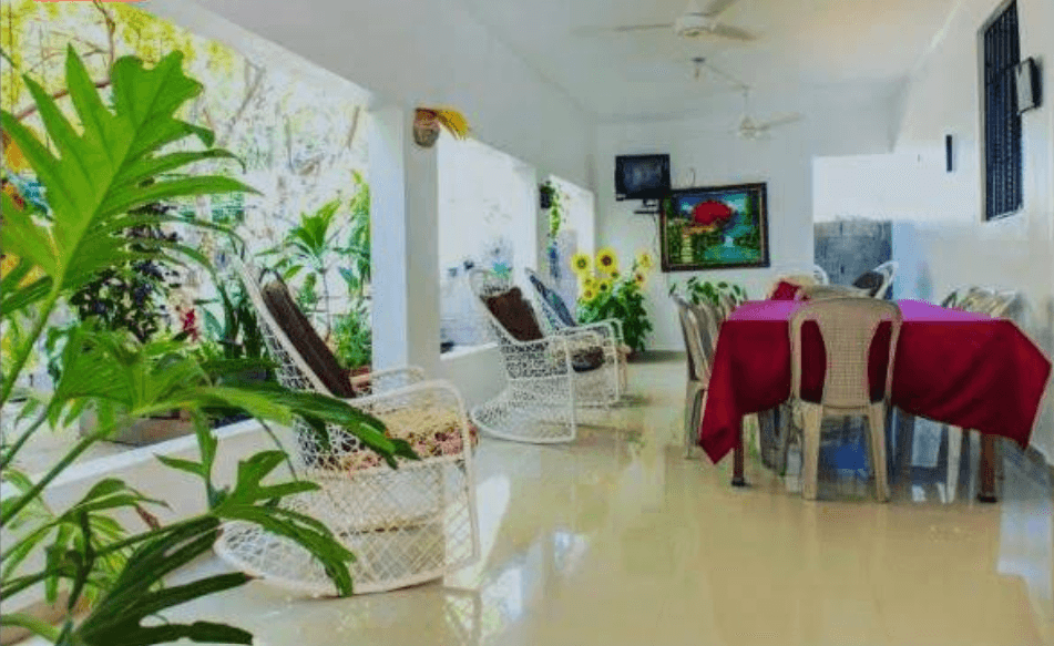 Terraza luminosa con mecedoras blancas, mesa con mantel rojo y exuberantes plantas verdes decorativas.